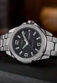Montre-bracelet en acier inoxydable argenté avec un cadran noir, index horaires lumineux, affichage de la date, et finition brossée sur la lunette et le bracelet.