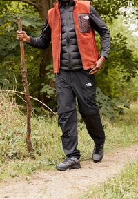 Botas de senderismo negras, pantalones negros con bolsillos con cremallera, chaqueta aislante negra, chaleco de forro polar naranja. Bastón de madera, fondo de follaje verde.