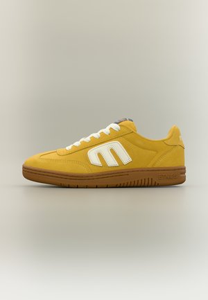 Baskets en daim jaune avec des lacets blancs et un logo, semelle marron portant la marque "Etnies", présentées de profil sur un fond neutre.