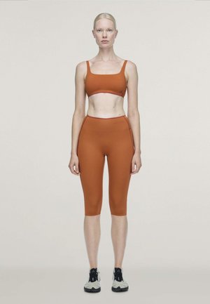 Un mannequin se tient de face, vêtu d’une brassière de sport et d’un short de cyclisme couleur rouille, avec des baskets grises, sur un fond uni.
