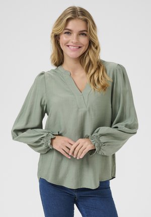 Blouse vert clair, texturée, avec un col en V, des manches bouffantes et des poignets froncés, associée à un jean bleu foncé. Design simple et décontracté.