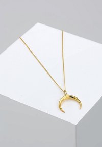 Pendentif en forme de croissant de lune en or suspendu à une fine chaîne en or, présenté sur une surface blanche inclinée sur un fond gris neutre.