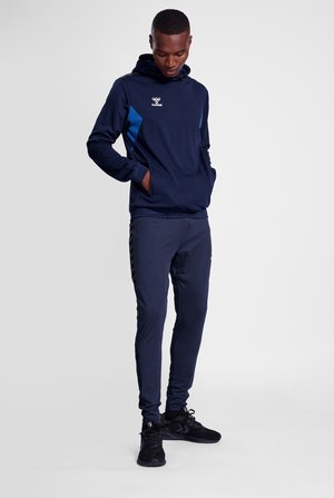 Marineblå sportshoodie og bukser med blå accenter. Hoodien har en kængurulomme og logo; bukserne har sidelister og en tætsiddende pasform.