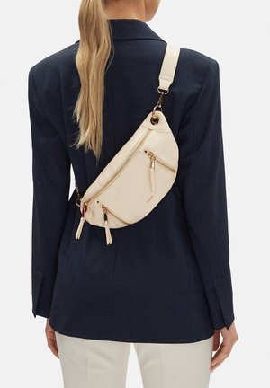 Mujer con cabello largo y rubio lleva una chaqueta azul marino oscuro y pantalones blancos, con un bolso bandolera color crema con cremalleras doradas en la espalda.