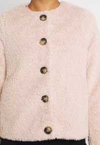 Lyserød, fluffet jakke med rund halsudskæring, der har fem store, mørke knapper og et tekstureret, blødt stof. Kort model med lange ærmer.