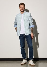 Košile na knoflíky v zelenobílé kostkované vzoru, vrstvená přes bílý tričko, spárovaná s tmavě modrými džíny a bílými teniskami.