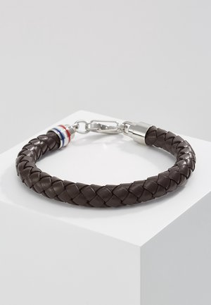 Flettet mørkebrun læderarmbånd med sølvspænde og rød, hvid og blå stribet ringdetalje, der hviler på en hvid piedestal.