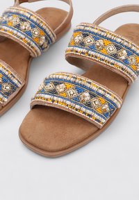 Sandalias de cuentas con tiras anchas que presentan adornos en azul, amarillo y plata sobre una base de ante color marrón claro. Suela plana y diseño de espalda abierta.