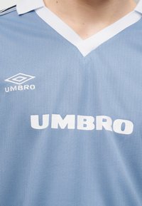 Μπλε αθλητική φανέλα με λευκό γιακά σε σχήμα V, με λευκό λογότυπο Umbro και εταιρική σήμανση στο στήθος.