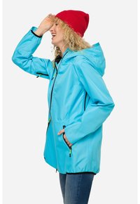 Giacca impermeabile azzurro chiaro con cappuccio, dotata di dettagli neri e tasche con zip, indossata con un berretto di lana rosso e jeans in denim chiaro.