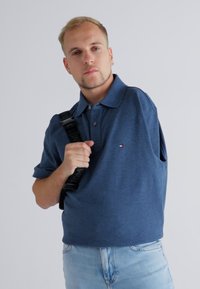 Kék póló, rövid ujjú, sima dizájnnal és kis logóval, világoskék farmernadrággal párosítva. A modell egy fekete pántos táskát tart.