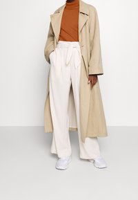 Trench-coat beige porté sur un col roulé rouille, associé à un pantalon blanc plissé taille haute et des baskets blanches, sur un fond uni.