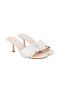 Mules en cuir blanc à bout ouvert, avec des clous en forme de pyramide dorés sur la sangle et un bout carré. Talon haut slim beige.