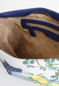 Väskan i vitt läder har blommigt mönster, blått foder, guldfärgad dragkedja och en ficka med dragkedja. Den är fodrad med beige tyg som har varumärkesnamnet tryckt på.