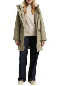 Langer olivgrüner Parka mit pelzbesetztem Kragen, getragen über einem cremefarbenen Strickpullover und dunklen Jeans, vervollständigt mit beigen Slippern.