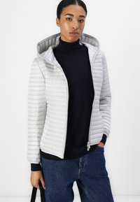 Gepolsterte, hellgraue Zip-Jacke mit Kapuze, mit horizontalen Nähten, gerippten Bündchen und glatter Innenfutter. Getragen über einem dunklen Pullover.