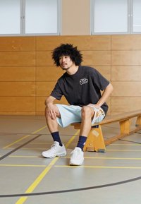 Camiseta negra de gran tamaño con logo, pantalones cortos azul claro con acentos florales, zapatillas blancas, sentado en un banco de madera en un gimnasio.
