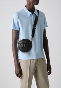 Light blue short-sleeve polo with zip collar, worn with beige trousers and a round black crossbody bag featuring embossed logo.
