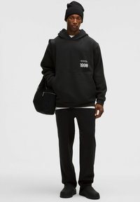 Czarny hoodie z przednią kieszenią, z białym graficznym napisem. Połączony z czarnymi dresami i gładkimi czarnymi butami wsuwanymi. Czarna czapka beanie.