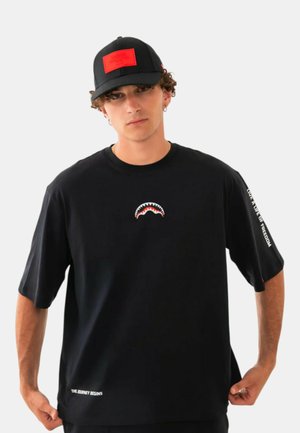 Giovane uomo che indossa un cappellino nero con toppa rossa e una T-shirt nera con grafica bocca di squalo e testo bianco sulla manica e sull'orlo inferiore.