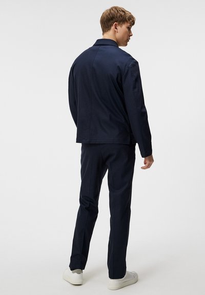 Marinblå kavaj och matchande byxor är tillverkade av ett mjukt tyg med en skräddarsydd passform. Lättviktsdesign, minimalistisk stil. Vita sneakers fulländar looken.