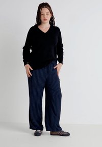 Suéter preto de decote em V combinado com calças largas, riscas azul-marinho. O tecido parece suave; os sapatos são escuros com riscas horizontais claras.