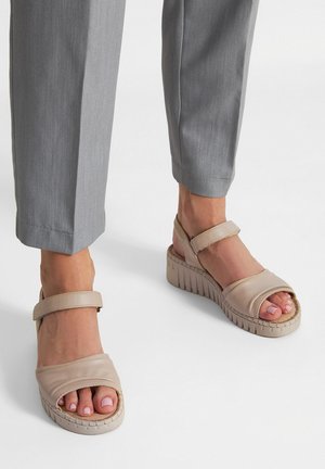 Füße in beigefarbenen Plateausandalen mit Knöchelriemen, kombiniert mit grauen, maßgeschneiderten Hosen vor weißem Hintergrund.