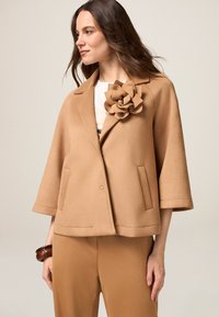 Blazer de lana color beige con un acento floral, mangas anchas y un corte relajado. Cuenta con dos bolsillos frontales y un cuello con muesca.