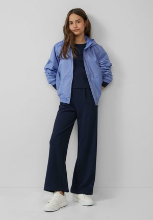 Lichtblauwe nylon bomberjack met capuchon, gecombineerd met een marineblauwe top en brede marineblauwe broek. Witte sneakers maken de outfit compleet.