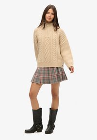 Pull beige en tricot avec un col montant et un motif en côtes, associé à une jupe plissée à carreaux gris et des bottes noires hauteur genou.