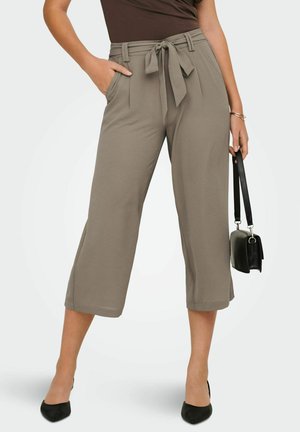 Femme portant un pantalon large court taupe avec ceinture nouée, haut noir, talons pointus noirs, tenant un petit sac à main noir.