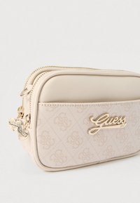 Bézs Guess csuklópánt arany logóval, mintás szövet elülső zsebbel és arany szív alakú cipzárhúzókkal világos háttéren.