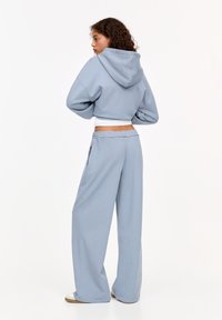 Haut de survêtement cropped bleu clair avec capuche, associé à un pantalon de survêtement ample. Tissu doux, semblable à un jersey, taille élastique et poches latérales.