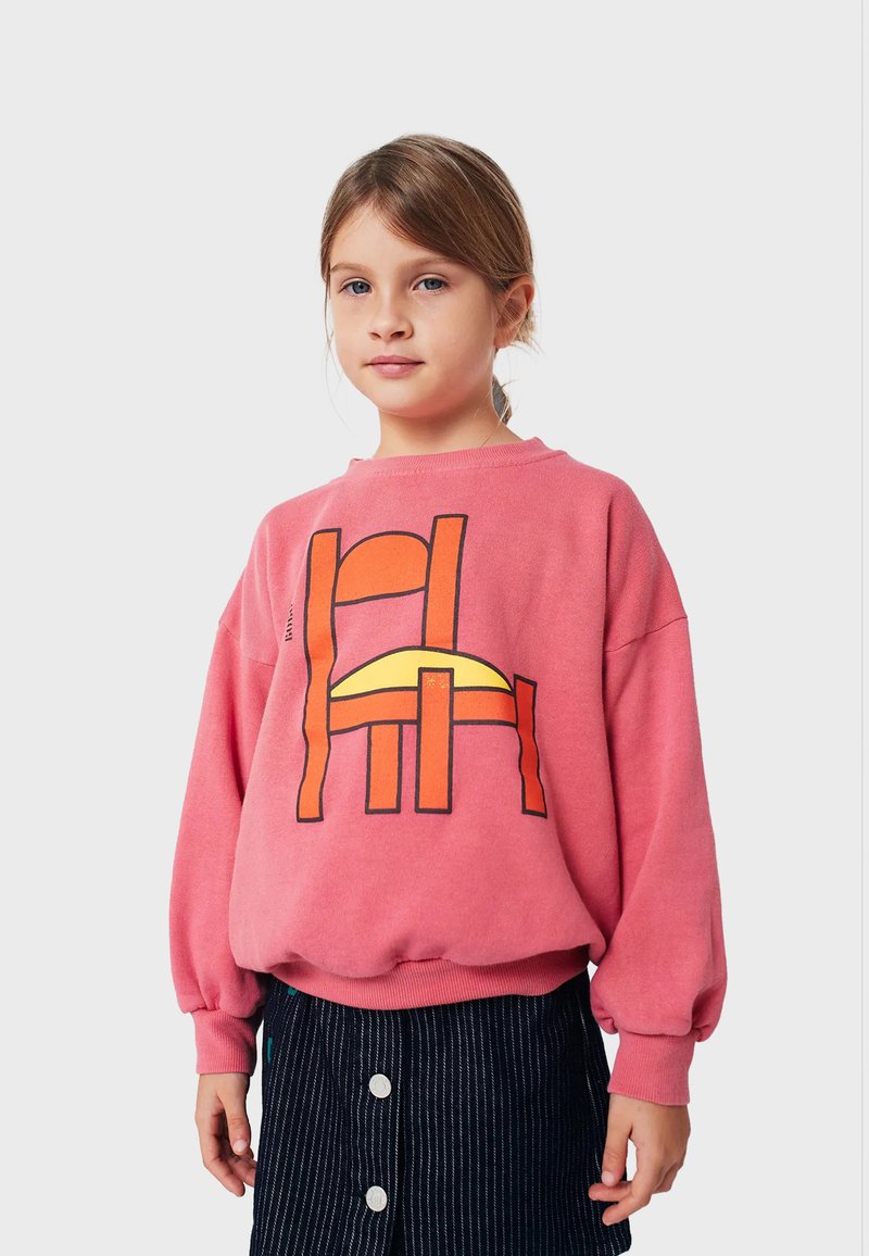 Felpa rosa con un design grafico featuring forme arancioni e gialle. Maniche lunghe con polsini elasticizzati, indossata sopra una gonna a righe scure.
