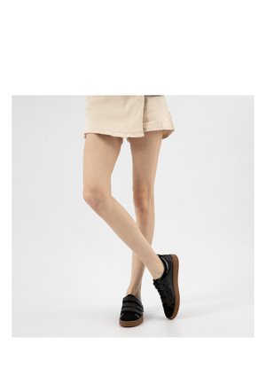 Osoba v béžových krátkých kalhotách a černých teniskách s hnědými podrážkami stojí se zkříženýma nohama na bílém pozadí.