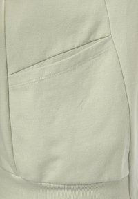 Hellgrüner Sweatshirtstoff mit weicher Textur, der über eine sichtbare schräge Tasche und ein geripptes Saumdetail verfügt. Nähte in kontrastierender Farbe.