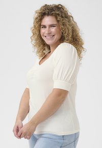 Top crème en matériau doux, avec un décolleté en V et de courtes manches bouffantes. La coupe est ajustée, mettant en valeur la silhouette.