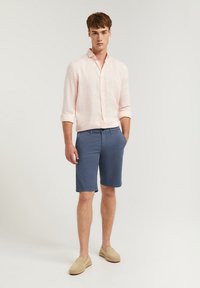 Chemise en lin rose clair, manches retroussées, shorts bleus, espadrilles beiges. Look décontracté, texture douce, sans motifs ni accents visibles.