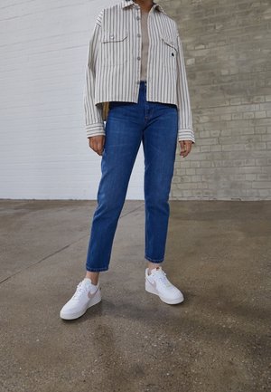 Osoba ubrana w pasiastą koszulę na guziki, beżowy top, niebieskie dżinsy i białe sneakersy Nike stoi na betonowej podłodze, za nią ceglany mur.