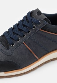 Marineblauwe sneakers van gestructureerd leer en mesh, met marineblauwe veters, bruine accenten en een lichtgekleurde zool.