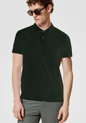 Uomo che indossa una polo verde scuro, pantaloni grigi e occhiali da sole, guarda verso il basso e leggermente di lato, su uno sfondo neutro.