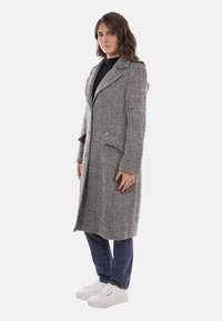 Cappotto grigio a motivo spina di pesce con colletto a incavo, tasche laterali e chiusura con un bottone, indossato sopra una maglia nera e jeans blu.