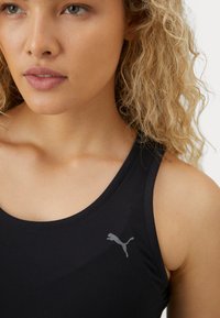 Camiseta de tirantes negra de deporte hecha de un tejido suave. Presenta un cuello redondo y un logo gris de Puma impreso en el pecho. Textura lisa.