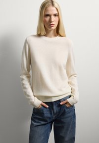 Crème kleurige trui van zacht breiwerk, met een ronde halslijn en ribbels aan de mouwen, gecombineerd met losse blauwe spijkerjeans.