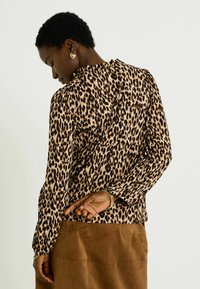 Top de manga larga con estampado de leopardo en tejido beige y negro, con un escote con volantes y puños ajustados, combinado con una falda de ante marrón.