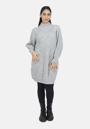 Vrouw draagt een grijze geribde gebreide sweaterjurk, zwarte leggings en zwarte vetercombatboots, staat met handen in de zij.