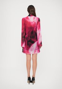 Uma mulher com cabelo longo e escuro veste um vestido mini rosa com flores e mangas longas transparentes, e sapatos de salto alto pretos, posando contra um fundo branco.