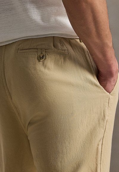 Κοντινό πλάνο του χεριού ενός ατόμου στην εμπρόσθια τσέπη μπεζ παντελονιού με κουμπωμένη πίσω τσέπη και λευκό πουκάμισο με υφή.