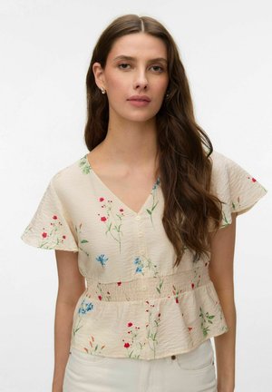 Kvinde med langt brunt hår iført kortærmet, cremefarvet bluse med blomsterprint og knapper, kombineret med hvide bukser foran en ensfarvet baggrund.