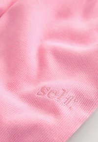 Ružová rebrovaná tkanina s jemnou textúrou, na ktorej je vyšívané logo "Self." Dizajn predstavuje čistú a jednoduchú estetiku.
